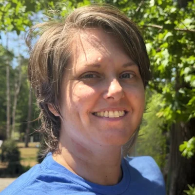 Smiling woman with short brown hair standing outdoors among green trees on a sunny day, wearing a blue shirt.