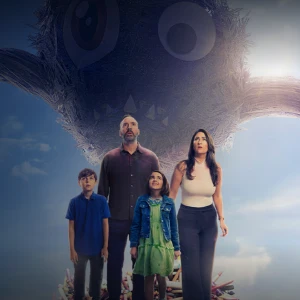 A family of four stands together outdoors as a huge creature's head looms in the blue sky behind them.