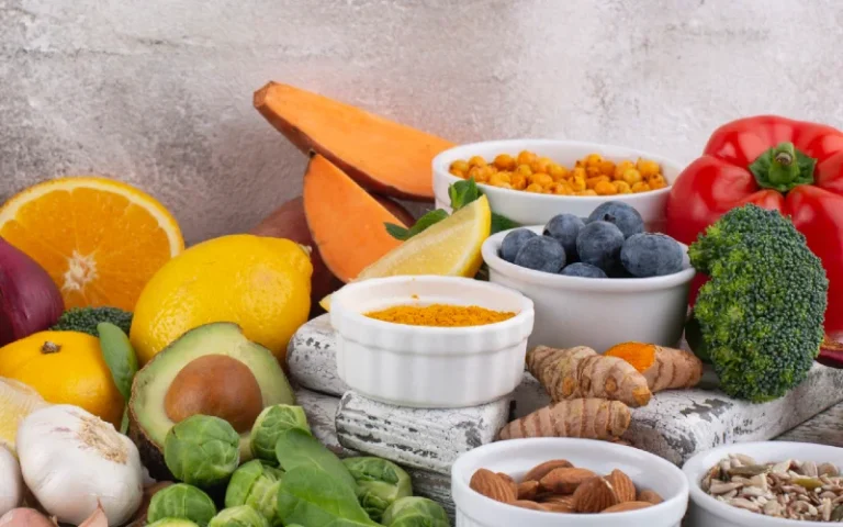 Assorted fresh fruits, vegetables, and nuts arranged on a table, featuring avocado, citrus, berries, broccoli, peppers, and spices in bowls
