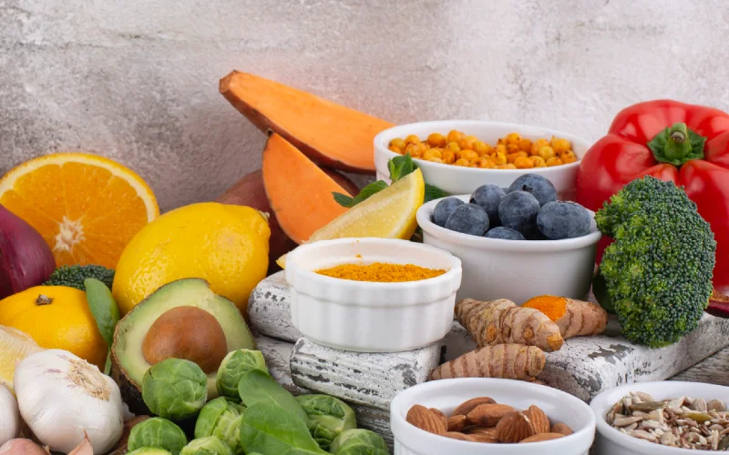 Assorted fresh fruits, vegetables, and nuts arranged on a table, featuring avocado, citrus, berries, broccoli, peppers, and spices in bowls