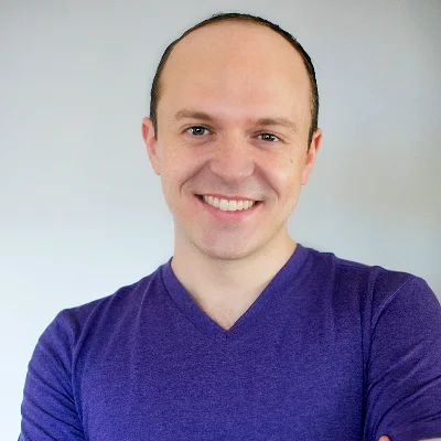 Portrait of a smiling man with short hair wearing a purple V-neck shirt, facing the camera against a light background.