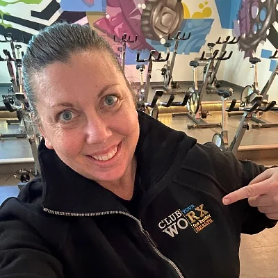 A woman takes a selfie in a gym with exercise bikes and a colorful mural in the background, wearing a dark zip-up hoodie with a small logo on the chest.