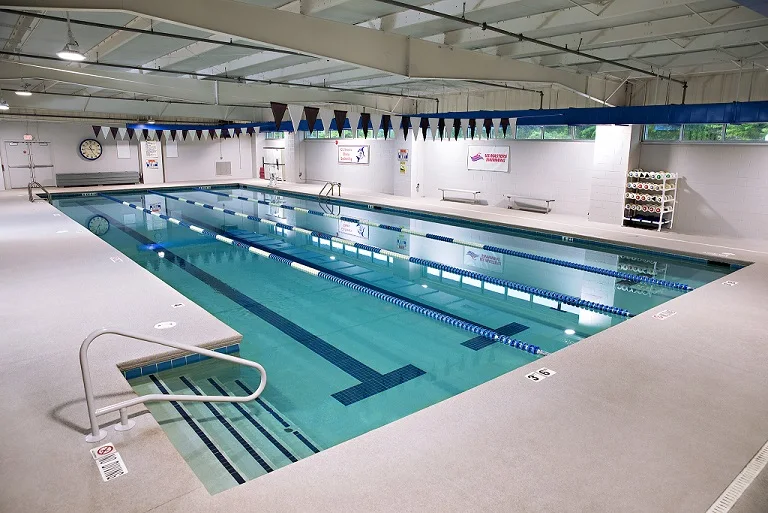 Indoor public swimming pool with three lanes, blue lane dividers, and a stair rail at the near corner of the deck while empty of swimmers.
