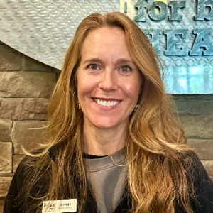 Smiling woman with long blonde hair wearing a name badge, indoors with a stone wall and teal artwork behind her.