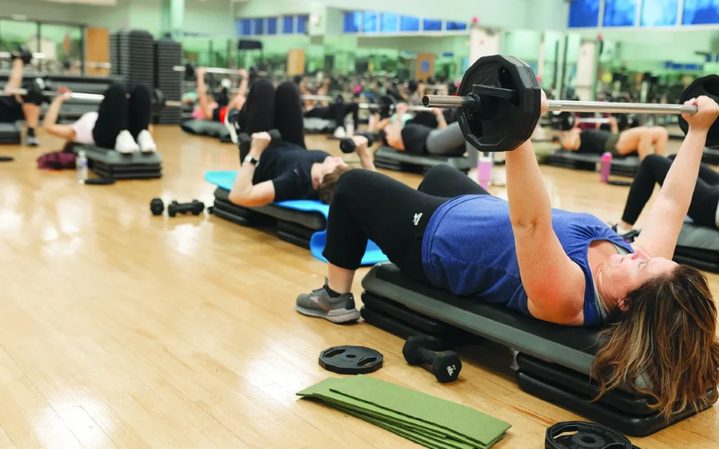Group fitness class in a gym: participants lie on mats on benches and press dumbbells overhead.