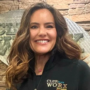 Smiling woman with wavy brown hair in a black jacket featuring a Club Worx logo, standing in front of a stone wall backdrop.