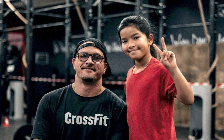 Man in CrossFit shirt and a girl in red posing in a gym, both smiling; girl shows peace sign at camera.