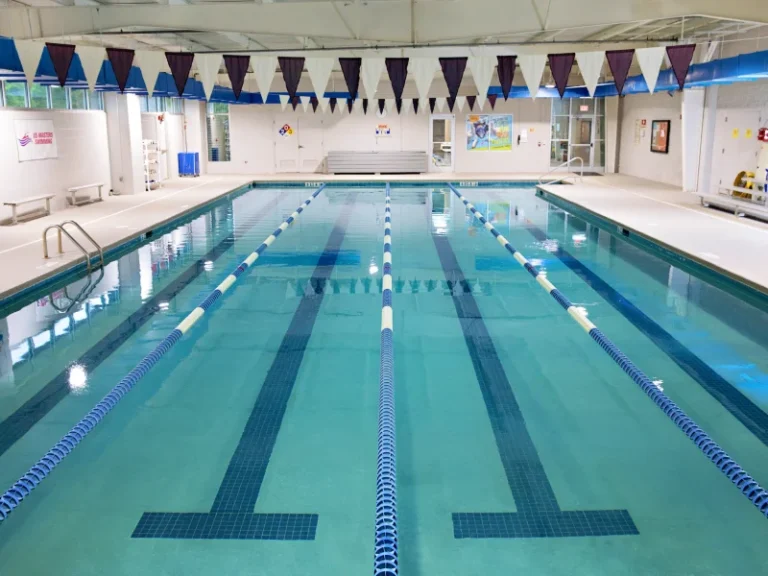 Indoor swimming pool with several blue lane lines, flags hanging overhead, and benches along the wall. The water is calm and reflective.
