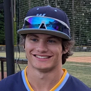 Young man wearing a navy baseball cap with mirrored blue sunglasses on top, smiling at the camera on a baseball field with a fence in the background.