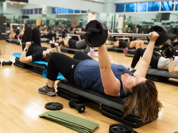 Person performing a bench press with a barbell on a padded bench in a gym, surrounded by others exercising on mats in the background