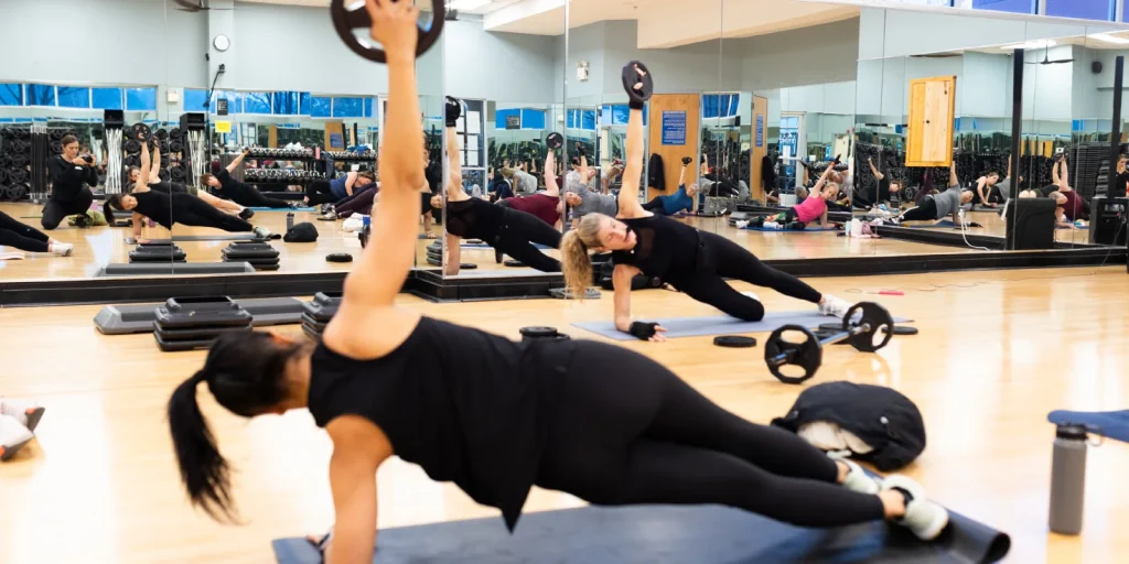 Group fitness class in a gym: participants on mats performing weighted side planks with dumbbells, mirrors and equipment around them.