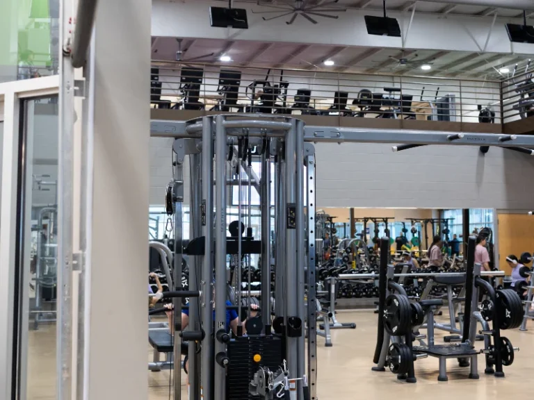 Gym interior with a cable weight machine in the foreground and other equipment and people exercising in the background.
