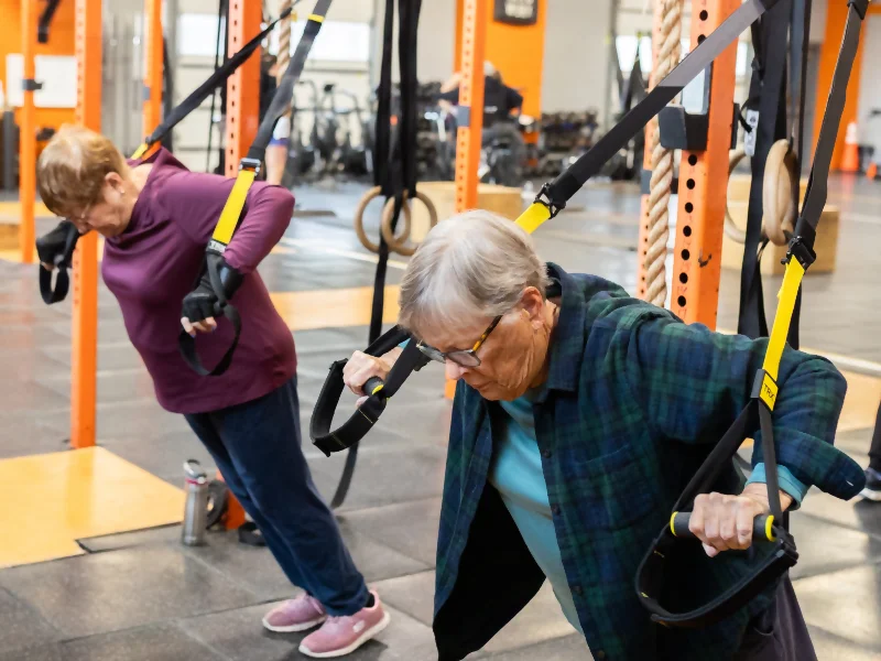 Two older women using suspension trainers in a gym, leaning forward and gripping the handles.