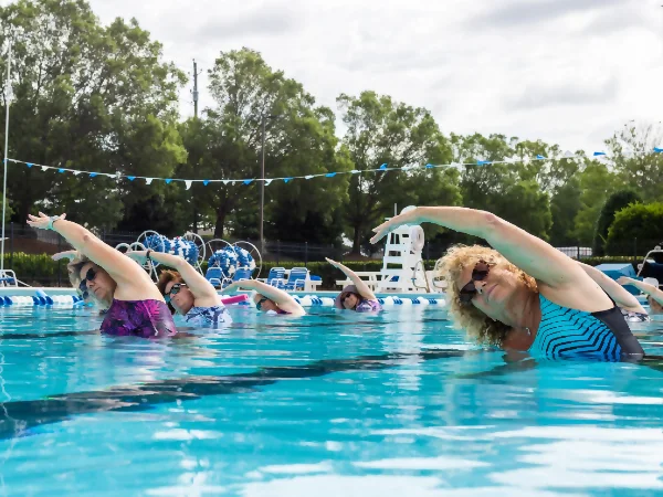 Group of adults doing water aerobics in an outdoor pool, bending to one side with arms overhead, wearing colorful swimsuits.