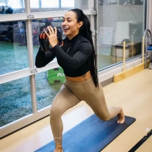 Woman in a black long-sleeve top and beige leggings performs a lunge with a medicine ball held at chest level inside a gym by a large window.