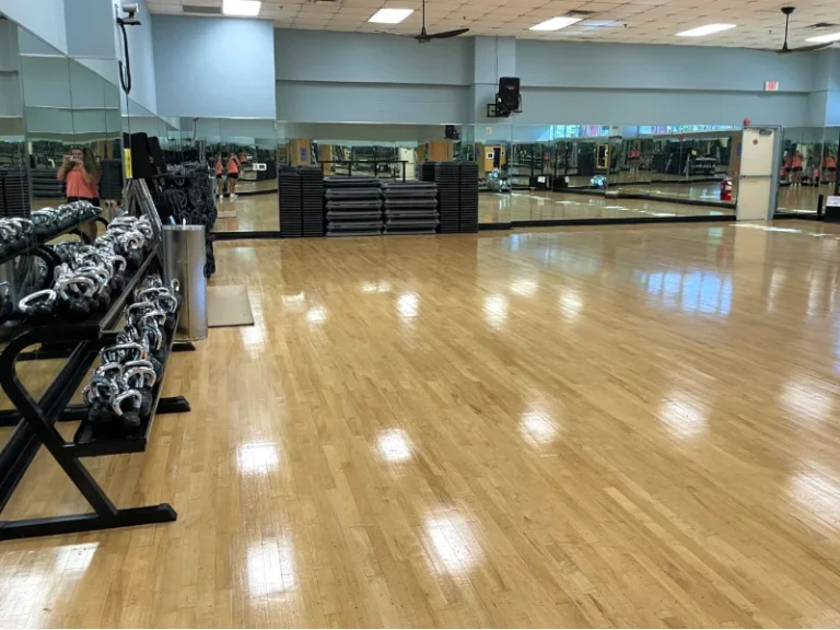 Empty fitness studio with a wooden floor and mirrored walls; dumbbells on a rack along the left side.