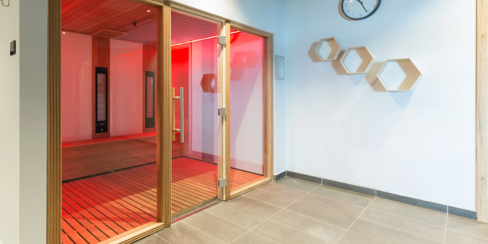 Glass sauna door entrance with red-lit interior; wooden panels and benches visible inside. A blue wall with hexagonal shelves and a clock on the right.