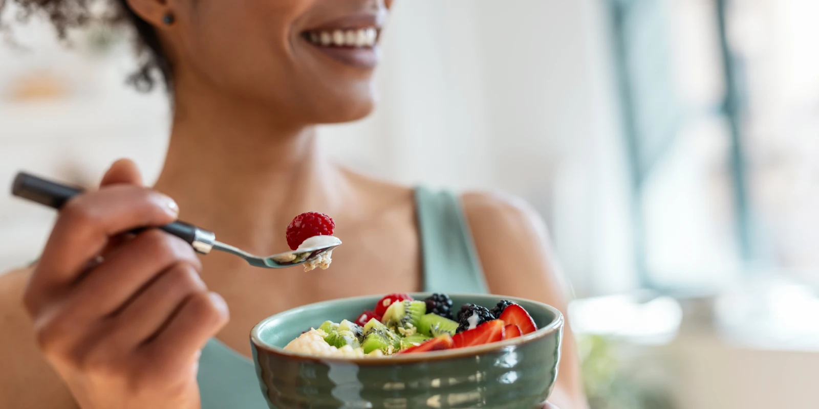 Smiling person holding a spoon with yogurt and a raspberry over a bowl of mixed fruit and yogurt parfait.