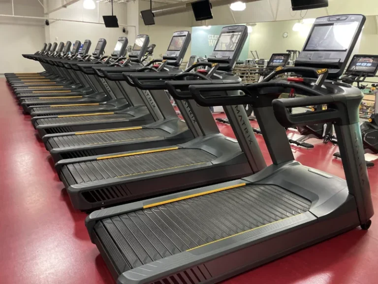 Row of treadmills lined up in a gym with large screens and red flooring behind them.