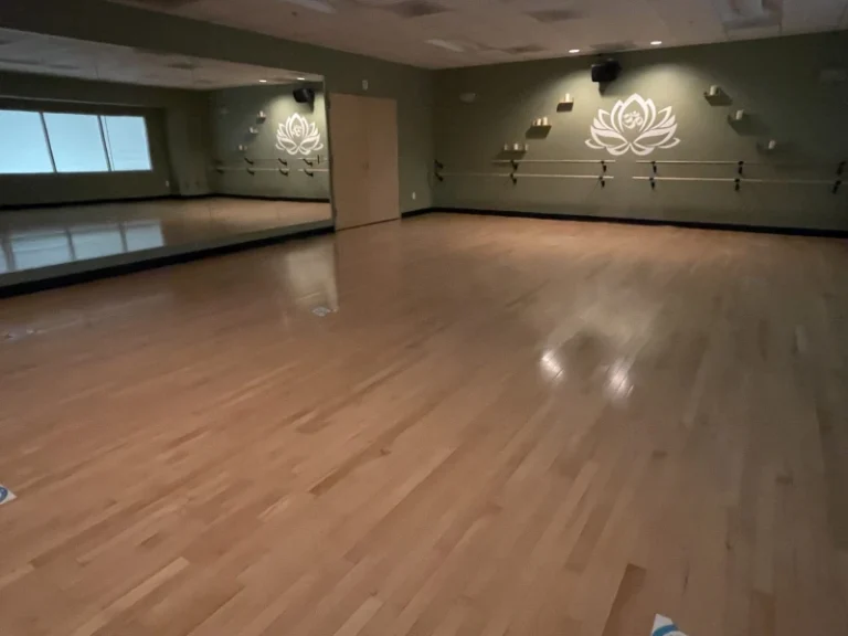 Empty dance studio with a wooden floor, large wall mirrors, ballet barre along the back wall, and a lotus logo on the green wall
