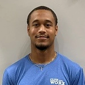 Portrait of a man with short hair and a light beard, wearing a blue t-shirt and a necklace, standing against a light gray wall.