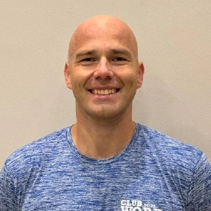 Smiling bald man in a blue athletic shirt standing against a beige wall.
